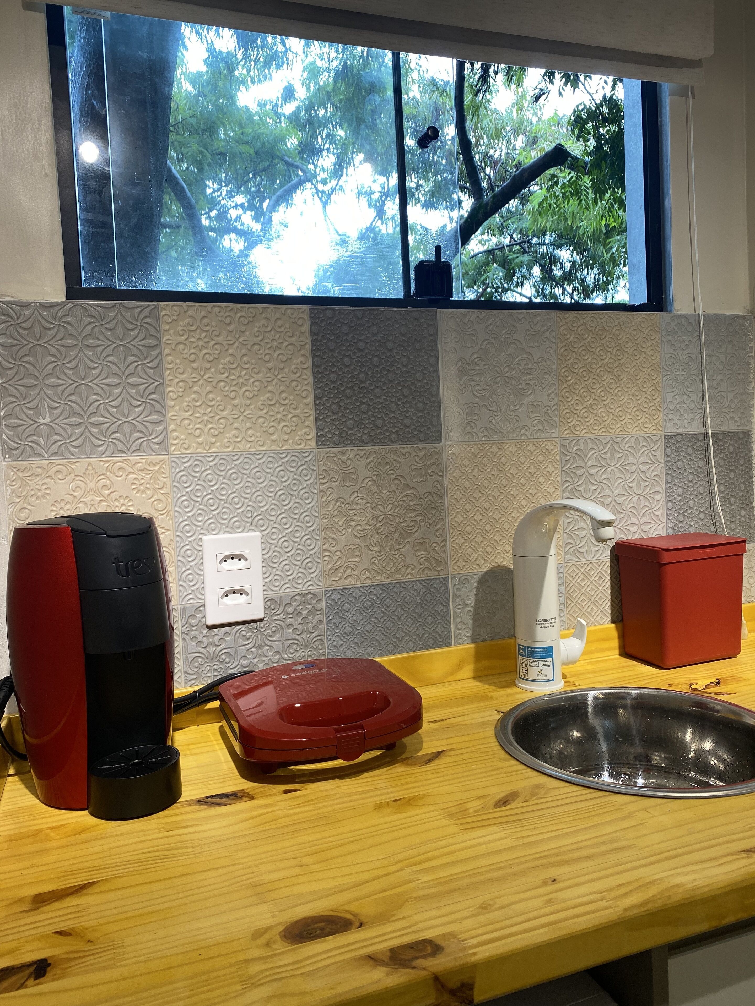 Een keuken met een raam, patroon betegelde vloer, een rode koffiezetapparaat, een rode container, een spoelbak en een witte zeepdispenser.