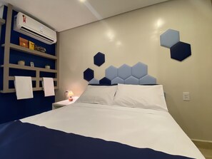 A bedroom with a bed, a nightstand, a lamp, a wall-mounted air conditioner, and a shelf with towels and decorative items.