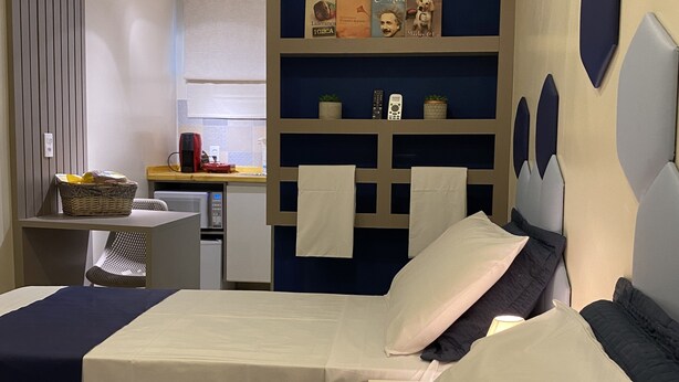 A hotel room with a bed, a desk, a chair, a shelf with decorative items, and an air conditioning unit. - Urban Box House (São Paulo)