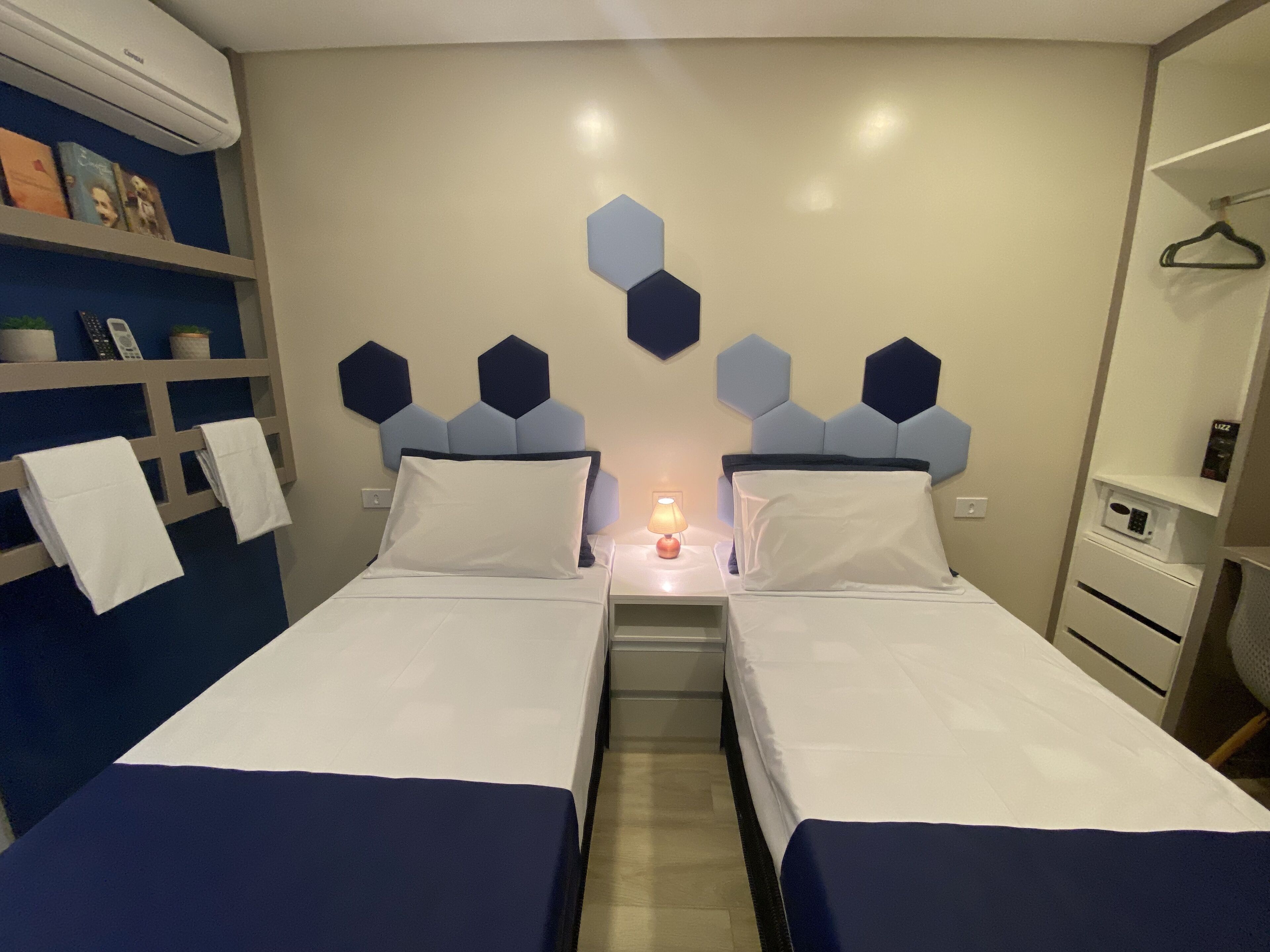 A hotel room with two beds, a bedside table with a lamp, a shelf with towels, and a wall-mounted air conditioner.