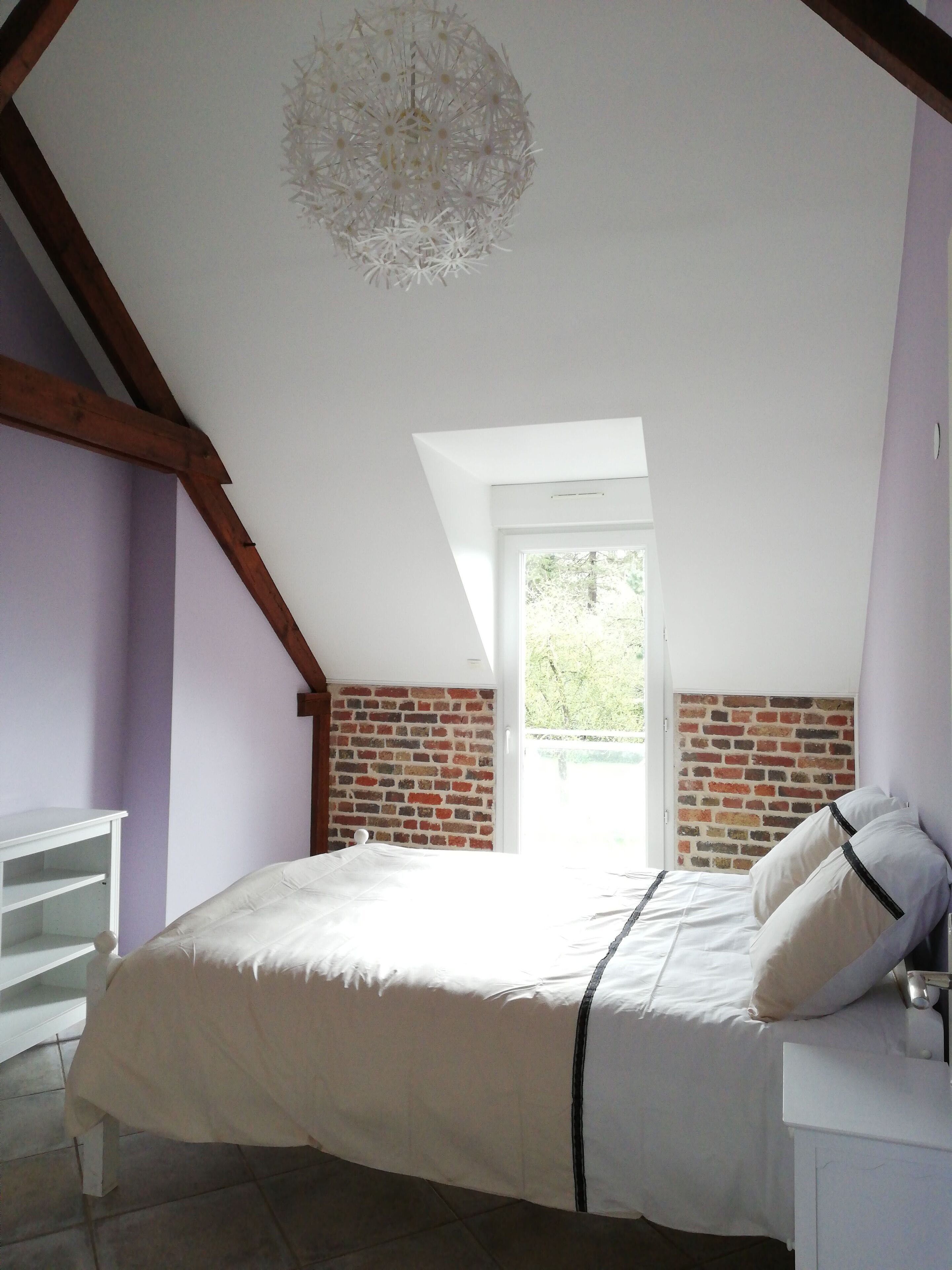 Une chambre avec un plafond en pente, un mur en briques, un lit avec une literie blanche et une fenêtre donnant sur un paysage verdoyant.