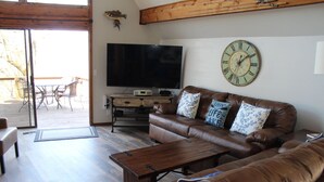 A living room with a leather sofa, a wooden coffee table, a flat-screen TV, and a wall clock. - Beautiful, Majestic Log Cabin Large Game Room and Lake View (Wagoner)