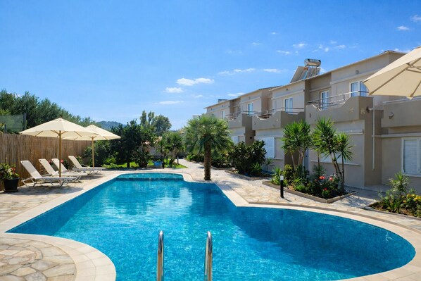 A swimming pool with lounge chairs and umbrellas, surrounded by greenery and buildings. - Secret Garden Hotel (Platanias)
