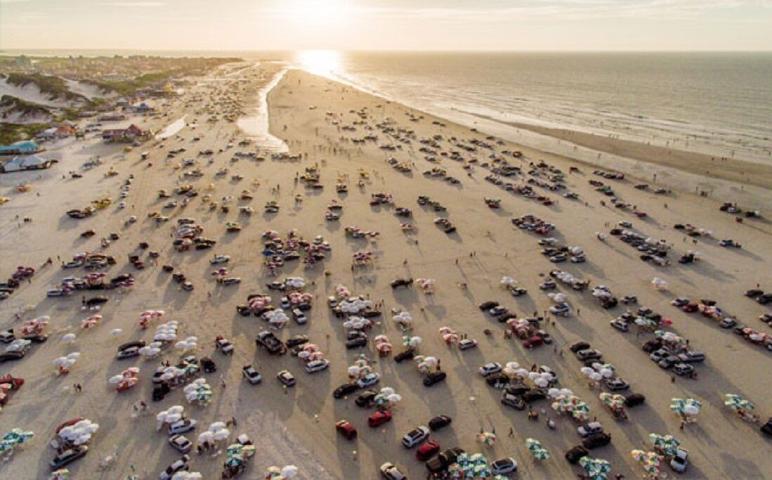 An aerial view of a beach with numerous parked vehicles and umbrellas.
