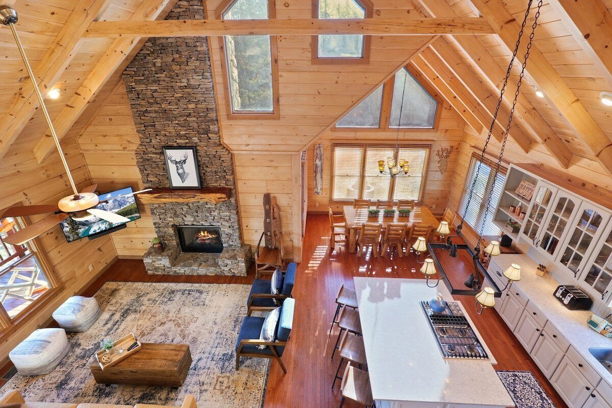 A spacious log cabin interior with a stone fireplace, wooden dining area, and large windows.