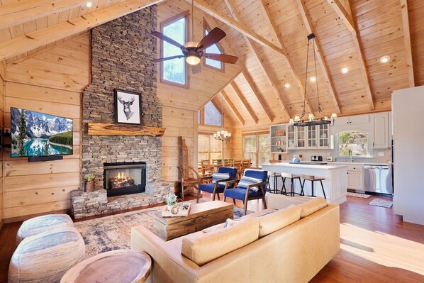 A spacious living area with a stone fireplace, wooden ceiling, and a kitchen in the background.