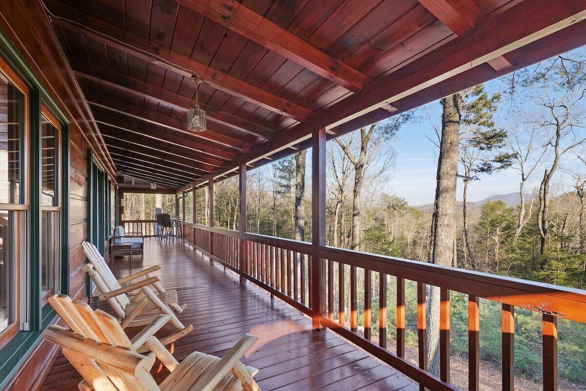 A covered porch with wooden railings, a seating area with chairs, and a view of a forested landscape.