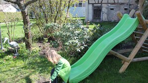A child playing near a green slide in a grassy area with trees and bushes. - Comfortable apartment on the Zollernalb for families, single travelers, workers, .... (Meßstetten)