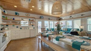A spacious kitchen with wooden cabinets, a dining area with a long table set for four, and a cozy seating area with a sofa and chairs. - Pool-Top Home-Walk to Town! (Port Aransas)