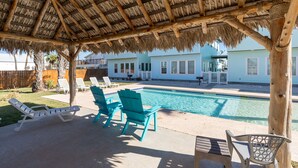 A pool area with a thatched roof, lounge chairs, and a view of a house in the background. - Pool-Top Home-Walk to Town! (Port Aransas)