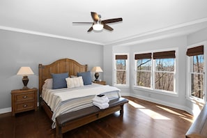 Un dormitorio con cama de madera, dos mesitas de noche con lámparas, un ventilador de techo y ventanas amplias con persianas.