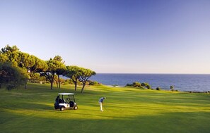 Um campo de golfe com um carrinho de golfe e um jogador se preparando para golpear a bola, rodeado por árvores e vista para o oceano.