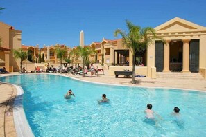 Piscina com pessoas na água, rodeada por palmeiras e edifícios de estilo mediterrâneo.