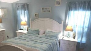 A bedroom with a bed, two nightstands, a window with curtains, and wall-mounted artwork. - Beachside Blessings (1 Block From the Beach!) (Mexico Beach)