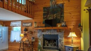 Interior acollidor d'una cabana amb una llar de pedra, parets de fusta, una televisió penjada i un sofà de cuir.