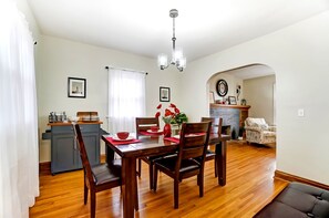 Un comedor con una mesa de madera, sillas y una araña. Se observa una zona de cocina con estufa y armarios.