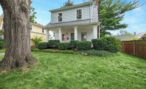 Une maison à deux étages avec un porche à l’avant, un grand arbre au premier plan et une pelouse bien entretenue.