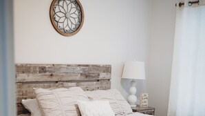 Un dormitorio con cabecera de madera, cama con ropa blanca, un adorno en la pared, una lámpara de noche y una mesita con un letrero.
