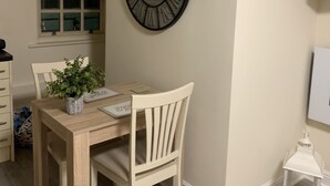 A dining area with a wooden table, chairs, a wall-mounted clock, a window with blinds, and a small white lantern. - 2-bed Duplex Sleeps 3 With Fireplace (Doncaster)