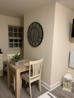 Un comedor con una mesa de madera, sillas, un reloj fijado a la pared, una ventana con persianas y un pequeño farol blanco.