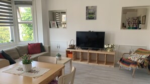 A living room with a sofa, TV, wooden table, chairs, and a window with blinds. - Spacious and bright modern styling 3 bed flat (LONDON)