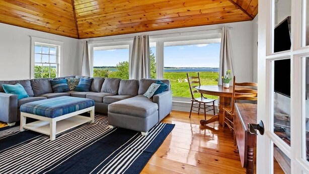 Ein gemütliches Wohnzimmer mit Holzbalkendecke, einem grauen Ecksofa, einem kleinen Tisch und Blick ins Freie.