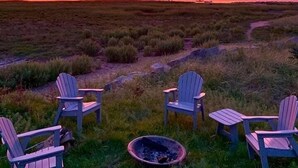 Ein Sonnenuntergang über einer grünen Landschaft, mit vier Adirondack-Stühlen, die um eine Feuerstelle angeordnet sind.