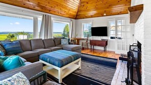 Ein gemütliches Wohnzimmer mit Holzboden, großen Fenstern, einer Couch, einem Couchtisch und einem Fernseher.