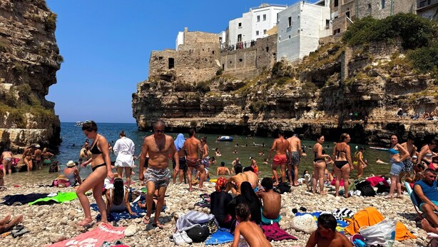 Une plage très fréquentée, avec des falaises rocheuses et des bâtiments blancs en arrière-plan.