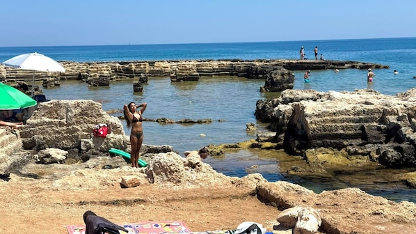 A rocky beach with clear blue water, people swimming and sunbathing, and a few umbrellas providing shade.