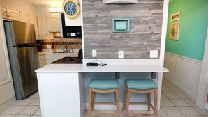 A kitchen with a stainless steel refrigerator, white cabinets, a wooden backsplash, two teal bar stools, and a clock on the wall.