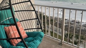 A balcony with a hanging chair, a patterned rug, and a view of the beach and ocean.