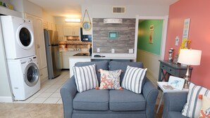 A compact living space with a grey sofa, a washing machine, a refrigerator, and a kitchen area with a wooden backsplash.