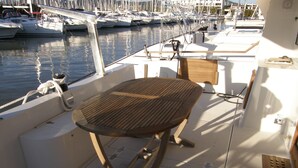 Ein Bootsdeck mit Holztisch, gepolstertem Sitz und Blick auf den Jachthafen mit weiteren Booten.