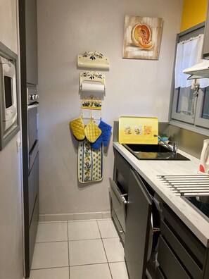 A kitchen with a white oven, a yellow sink, and a stainless steel range hood. There are hanging kitchen towels and a decorative wall piece above the oven. - Rent appt.studio T1 (29 m2 +loggia 7 m2) Rsd le Parc des Oliviers la FAVIERE (Bormes-les-Mimosas)