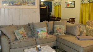 A cozy living room with a sectional sofa, a small white table, and patterned carpet. - Location Location Location! Classic Beach Cottage less than 1-min walk to beach! (Murrells Inlet)