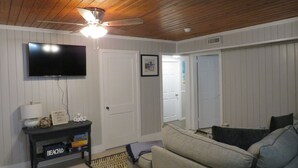 A living room with a ceiling fan, a flat-screen TV, a sofa, and a console table with a lamp and decorative items. - Location Location Location! Classic Beach Cottage less than 1-min walk to beach! (Murrells Inlet)