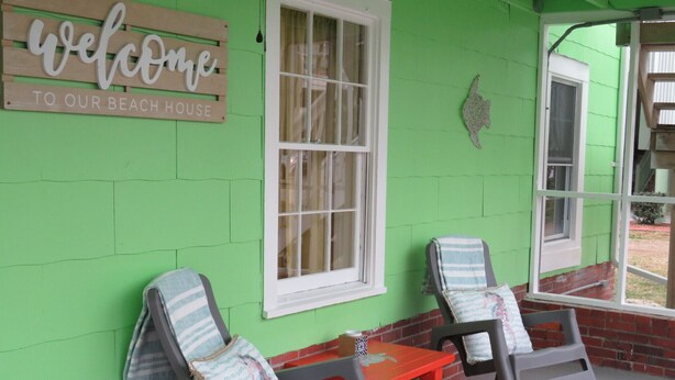 A beach house exterior with a 'Welcome to Our Beach House' sign, two rocking chairs, a small table, and a window. - Location Location Location! Classic Beach Cottage less than 1-min walk to beach! (Murrells Inlet)