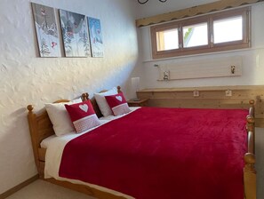 Quarto com cama de madeira, roupa de cama vermelha e branca, janela com persianas de madeira e arte pendurada na parede.