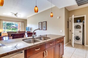 A kitchen with granite countertops, a double sink, wooden cabinets, pendant lighting, and a view of a living area with a ceiling fan.