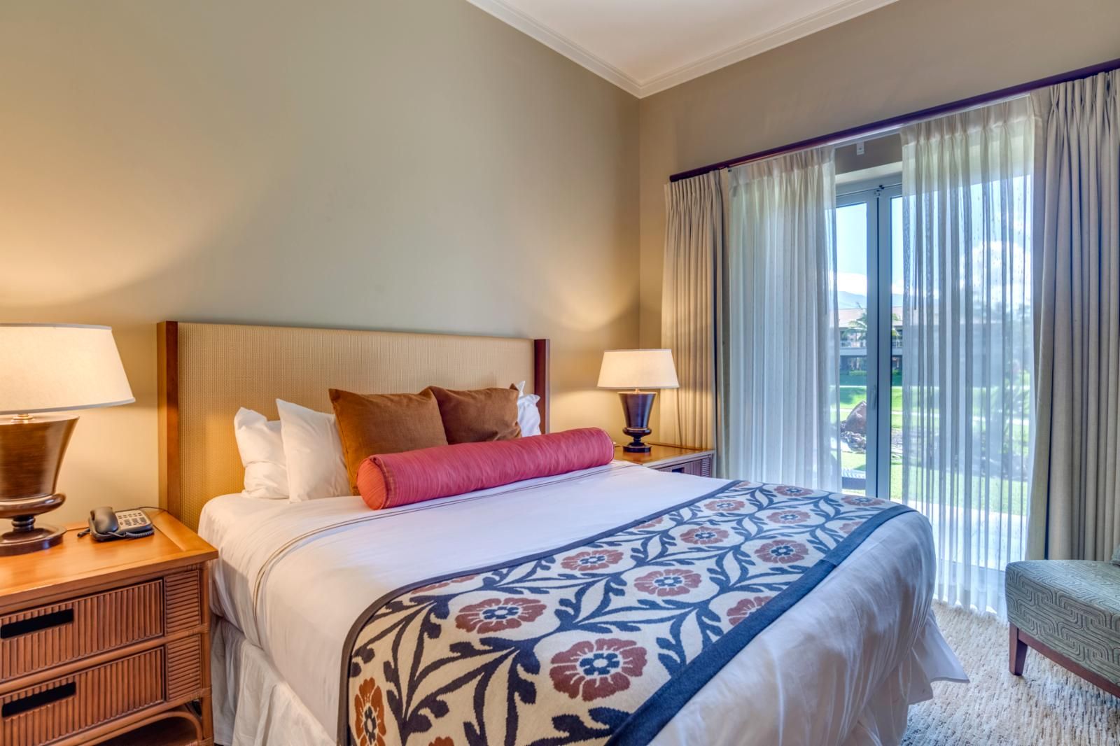Habitación de hotel con una cama grande, dos mesitas de noche con lámparas, una mesita auxiliar de madera y vistas al exterior a través de una ventana con cortinas.
