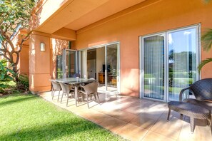 A patio with a glass table and chairs, a sliding glass door, and a view of a lawn and trees.