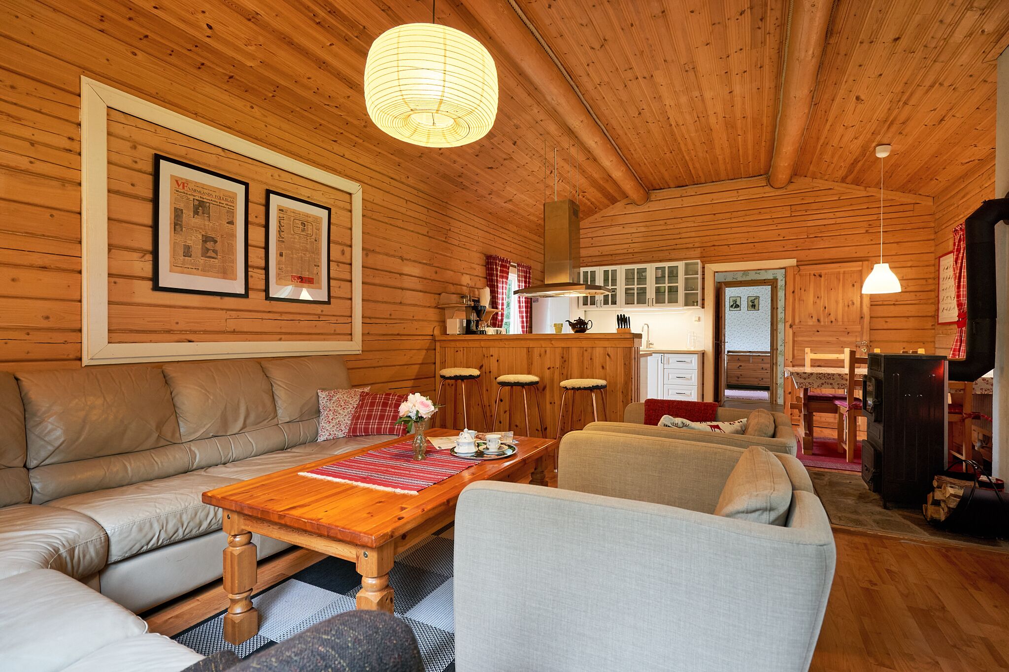 A cozy cabin interior with a wooden ceiling, a dining table with chairs, a sofa, and a kitchen area visible through an open door.