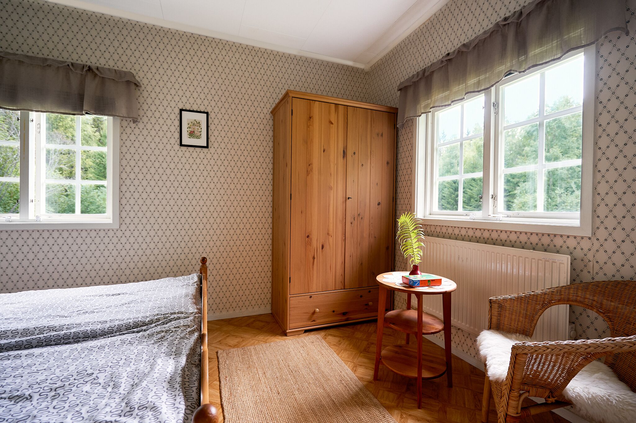 A room with a bed, a wooden wardrobe, a wicker chair, a small table with a plant, and patterned wallpaper.