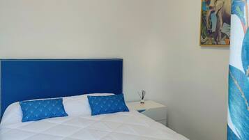Een bed met een blauw hoofdbord en witte beddengoed, een klein nachtkastje met een plant, en een muur met een poster met een tropisch thema.