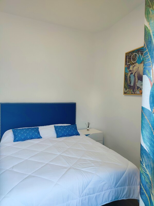 A bed with a blue headboard and white bedding, a small bedside table with a plant, and a wall with a tropical-themed poster.