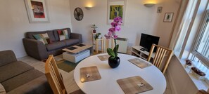 A modern living room with a grey sofa, a white coffee table, and a TV. There are two potted plants and a small table with a decorative item.