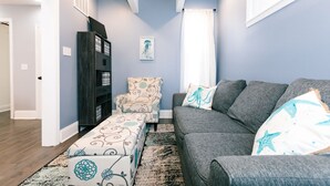 A living room with a grey sofa, patterned ottoman, and a dark wooden bookshelf. - 3-BR house walk to beach, shopping, bars, NYC train. 6-beach passes & 4-bikes. (Asbury Park)