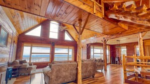 Interior espacioso de una cabaña de troncos con amplias ventanas, pisos de madera y una zona de estar acogedora.