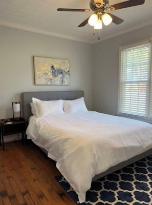 A bedroom with a bed, a nightstand, a ceiling fan, a window with blinds, and a wall-mounted painting.